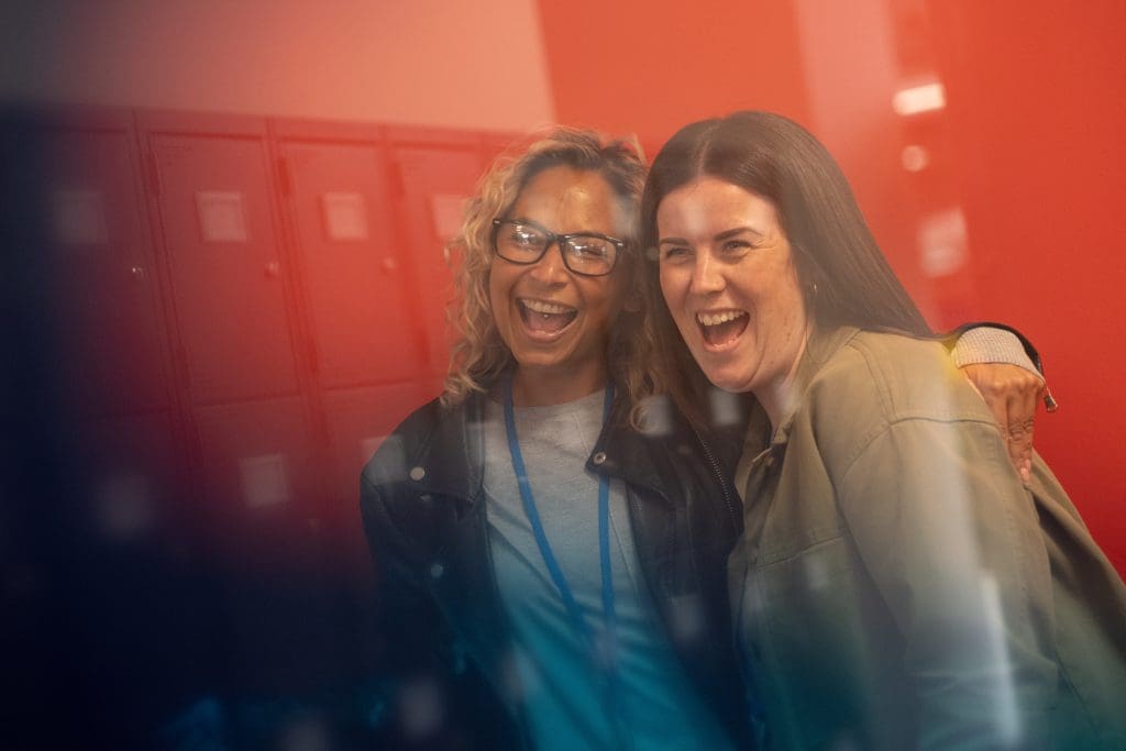 two women, one with arm around the other both smiling in locker room